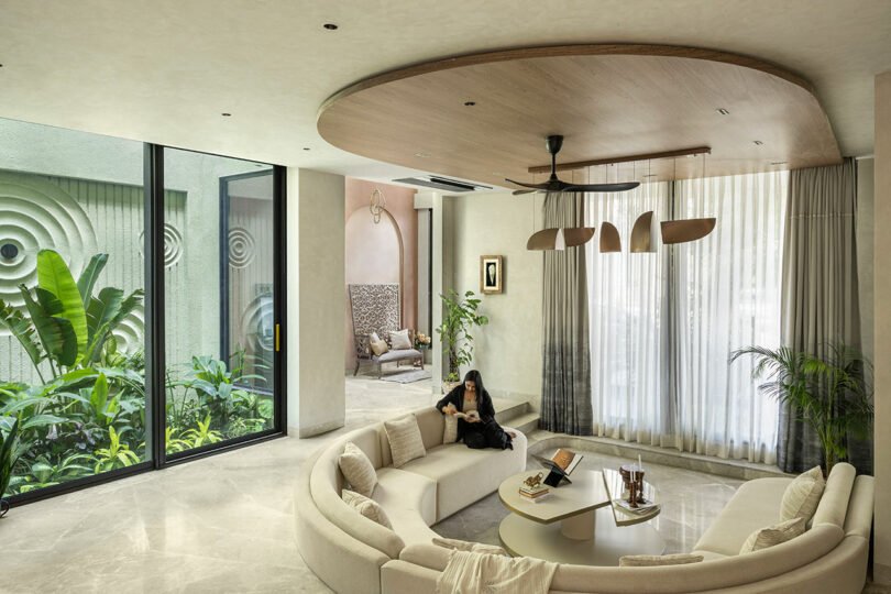 A woman sits on a curved beige sofa in a modern living room with large windows, indoor plants, and natural light.