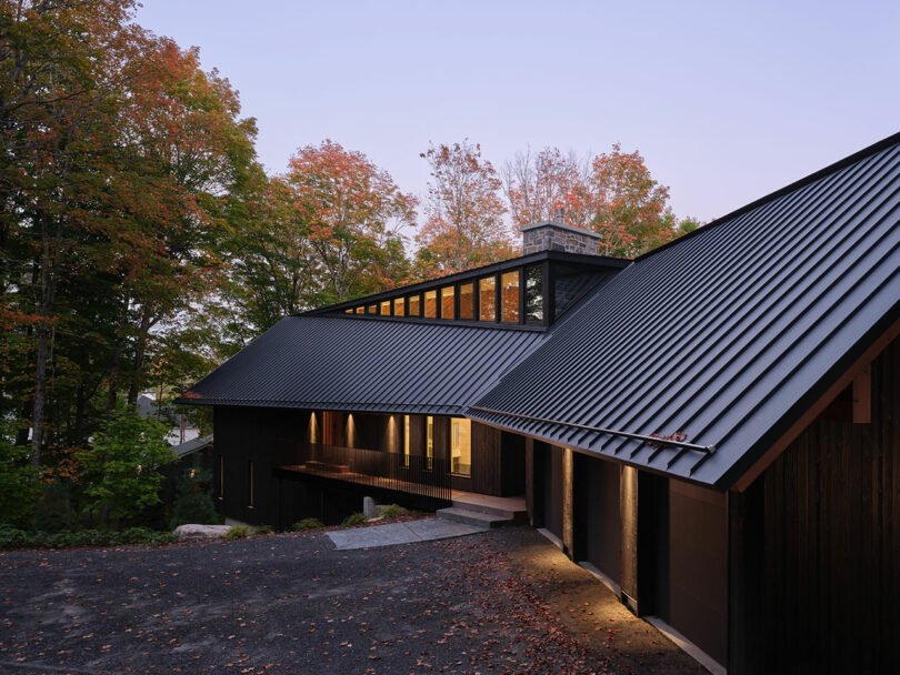 Modern house with dark metal roof and wood siding, surrounded by autumn trees at dusk, with warm interior lights visible through large windows.