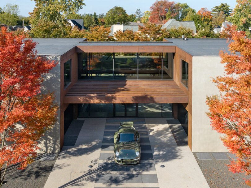 A green vintage car is parked in the driveway of a modern, two-storey house with large windows, surrounded by trees with autumn-coloured leaves.